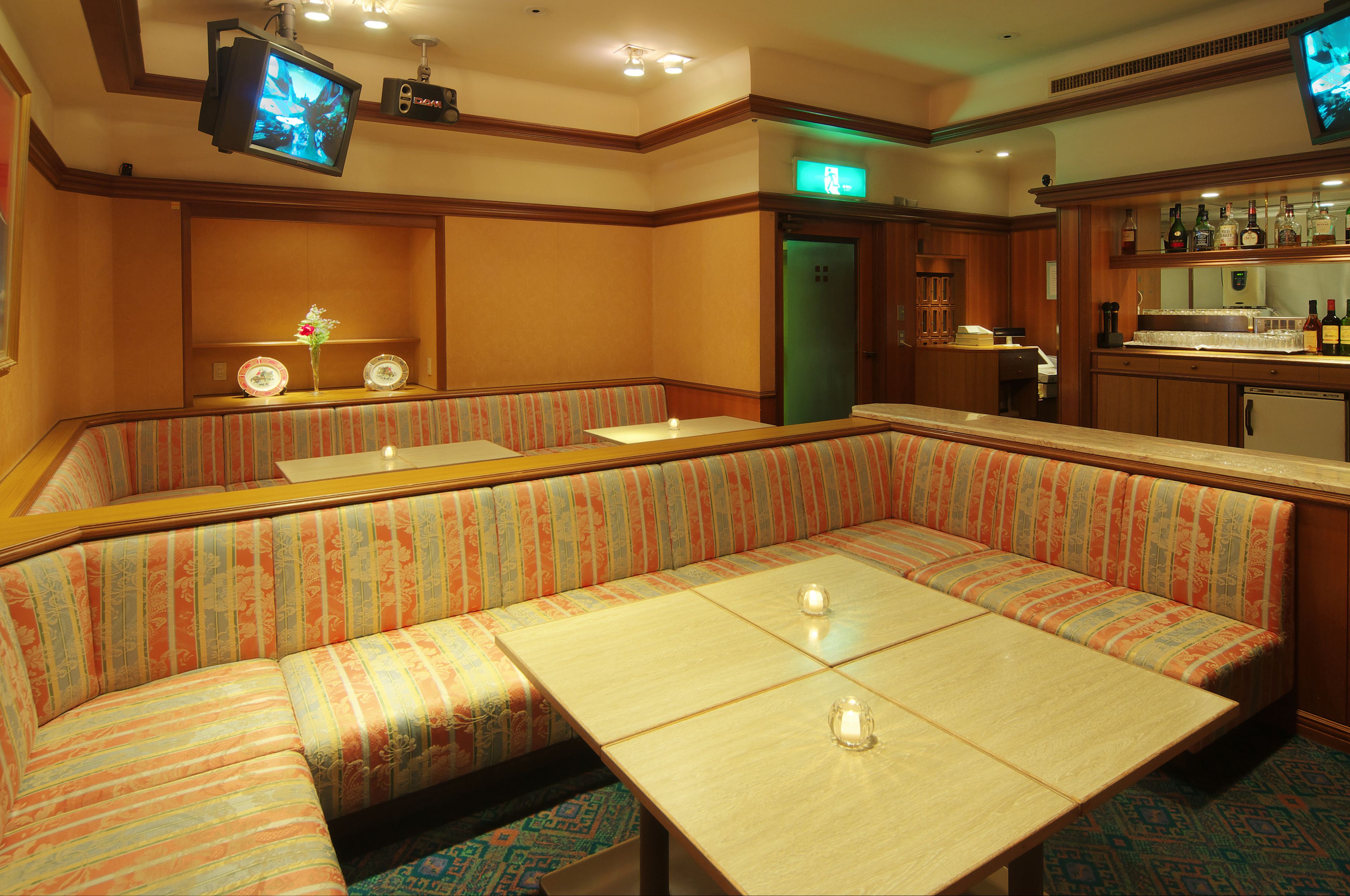 A hotel lobby with a large, striped booth seating area, a bar with a counter and shelves of bottles, and a television mounted on the wall.