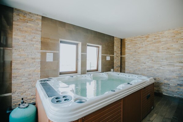 Un amplio jacuzzi interior con base de madera, frente a una pared de piedra con dos ventanas.