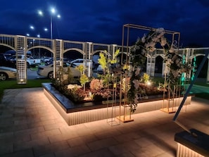 A paved outdoor area with a decorative metal structure and plants, surrounded by a fence and underlit by street lamps. - Hotel Romanita (Baia Mare)