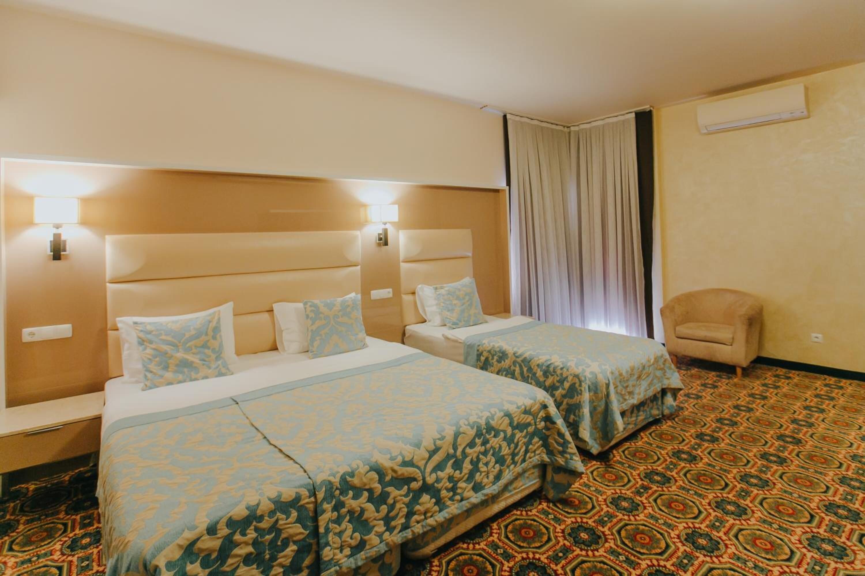 A hotel room with two beds, a chair, a patterned carpet, and a curtain.