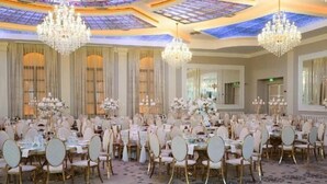 Un salón de banquetes con mesas redondas, sillas blancas, candelabros y una alfombra estampada.