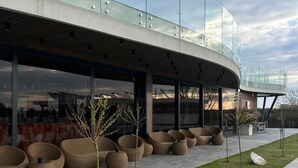 Un edificio moderno con fachada de vidrio curva, zona de estar exterior con sillas de mimbre y un sendero pavimentado.