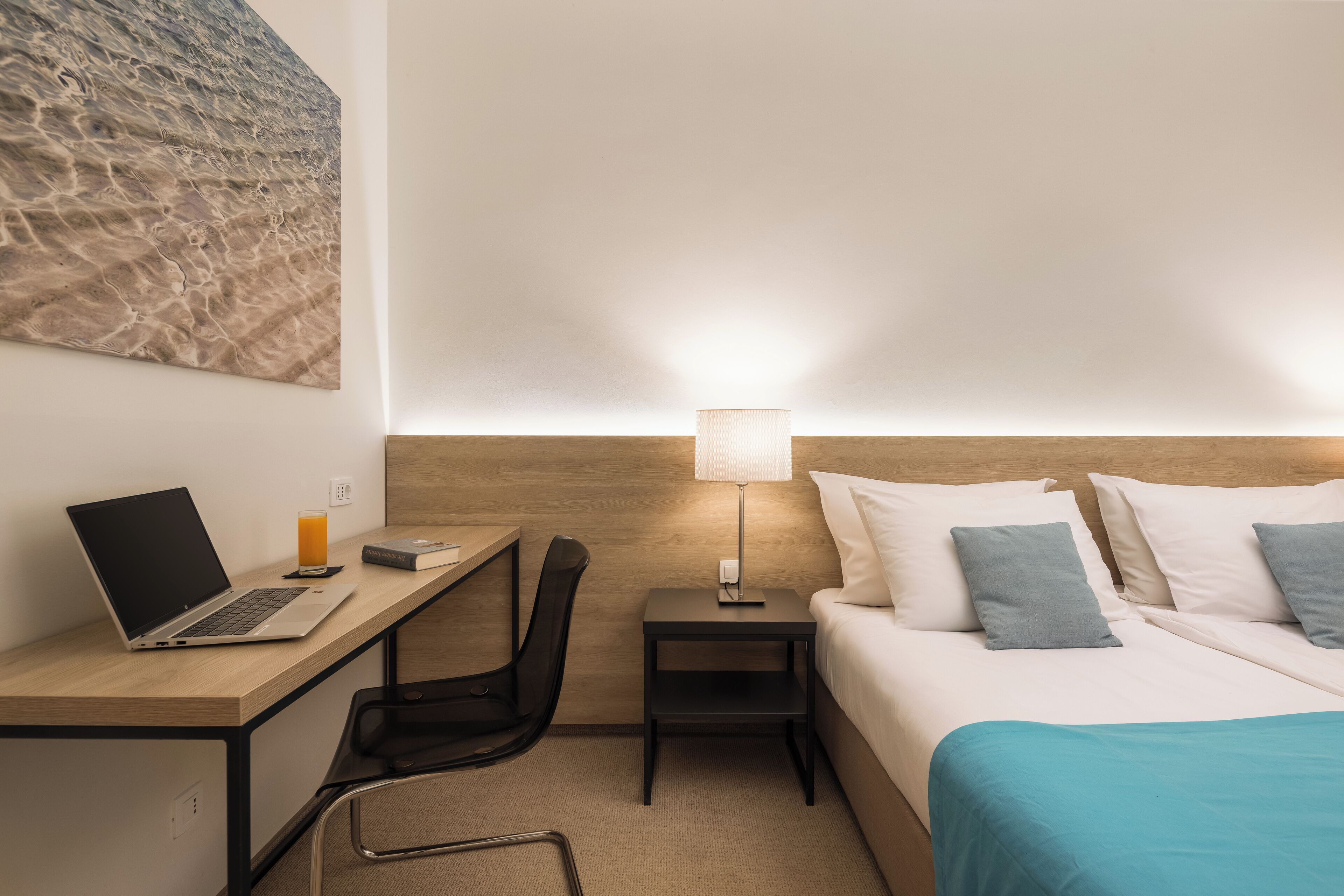 Une chambre d’hôtel comprenant un lit, un bureau avec un ordinateur portable, une chaise, une table de chevet avec une lampe et une œuvre d’art murale.