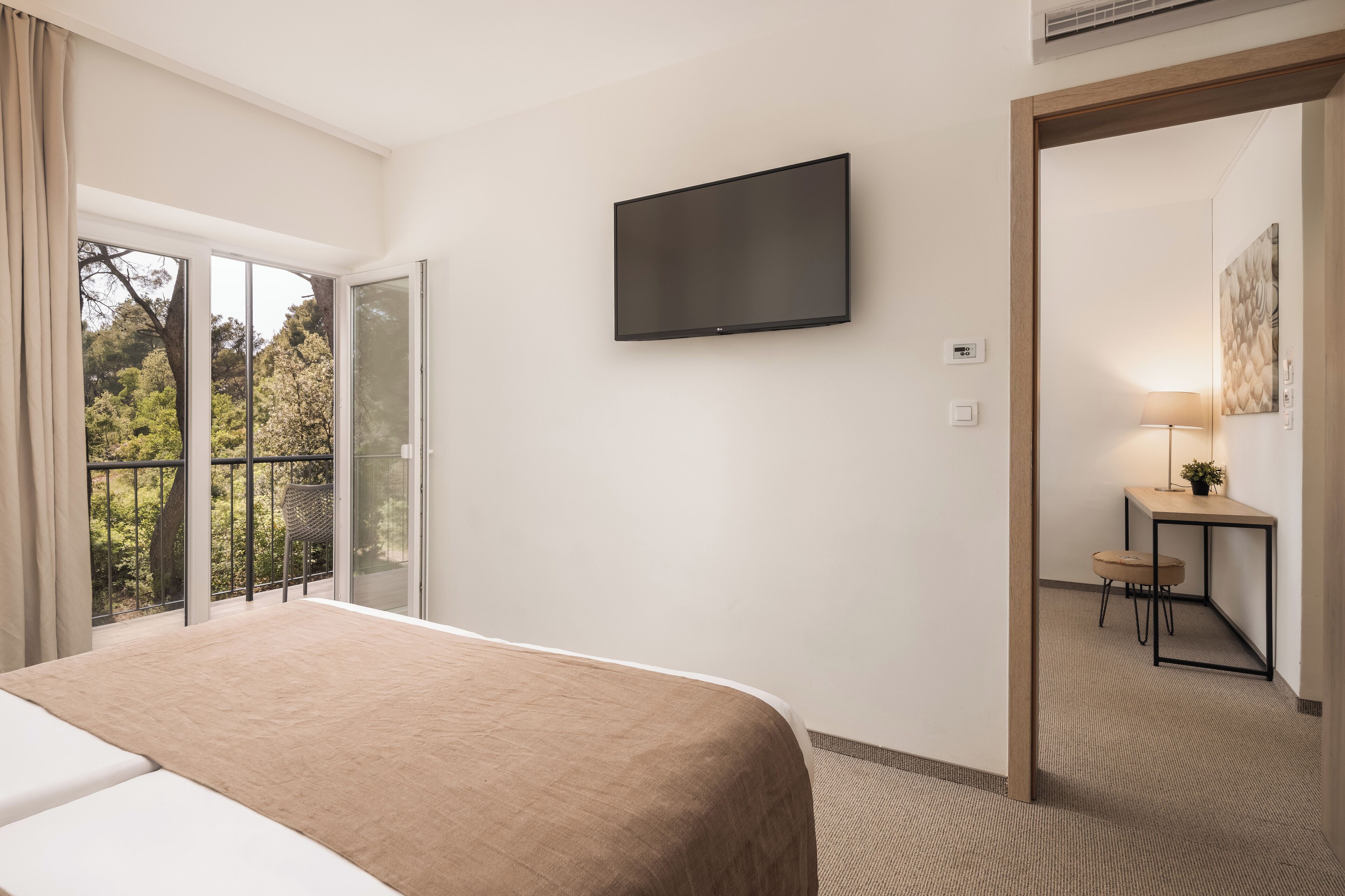 A hotel room with a bed, a TV mounted on the wall, a small desk with a chair, and a view of trees through the window.