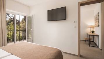 A hotel room with a bed, a TV mounted on the wall, a small desk with a chair, and a view of trees through the window.