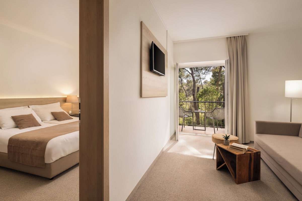 A hotel room with a bed, a sofa, a TV mounted on the wall, a small table with a lamp, and a view of the outdoors.