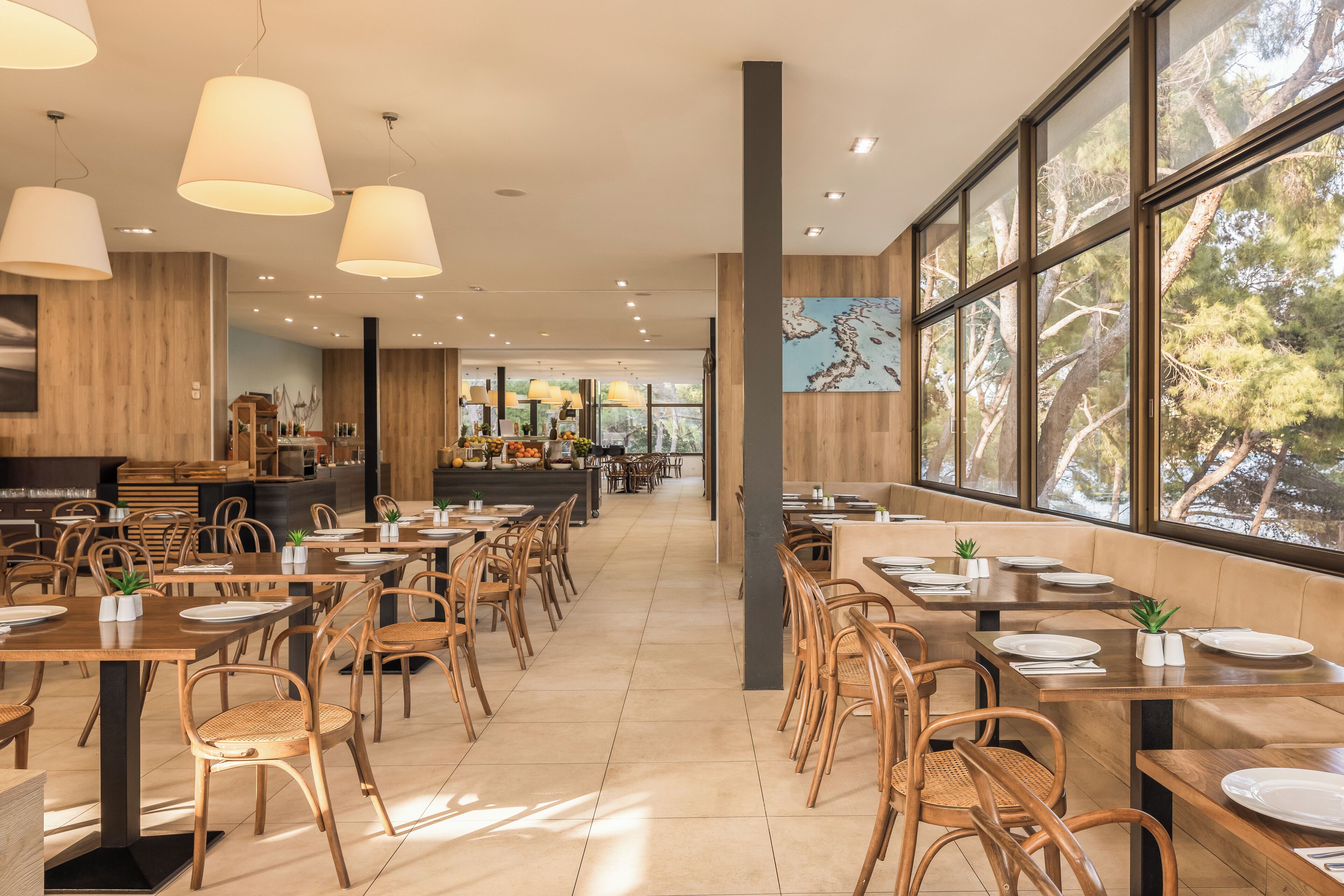 A spacious restaurant with wooden tables and chairs, large windows offering a view of trees, and pendant lighting.