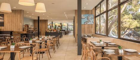 A spacious restaurant with wooden tables and chairs, large windows offering a view of trees, and pendant lighting.
