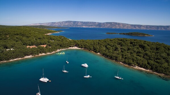 A coastal area with clear turquoise waters, several sailboats anchored near the shore, and a backdrop of dense green hills.