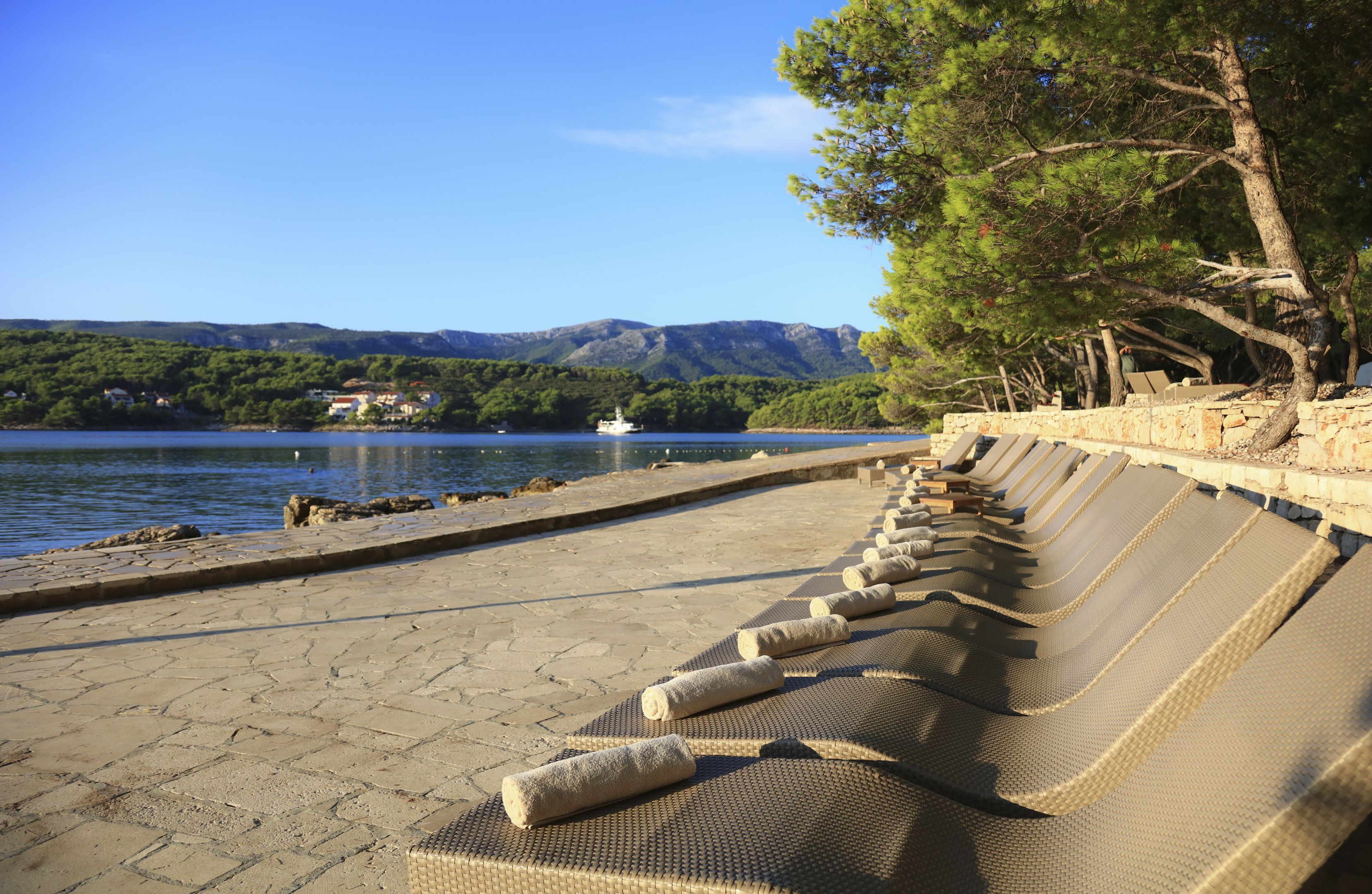 A row of lounge chairs with towels, a stone pathway, a body of water, and a boat in the distance.