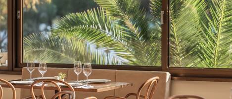 A dining area with a table set for two, wooden chairs, and a view of palm leaves through a window.