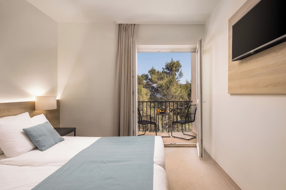 A hotel room with a bed, a nightstand, a TV mounted on the wall, a balcony with a table and chairs, and a view of trees and sky.