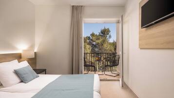 A hotel room with a bed, a nightstand, a TV mounted on the wall, a balcony with a table and chairs, and a view of trees and sky.