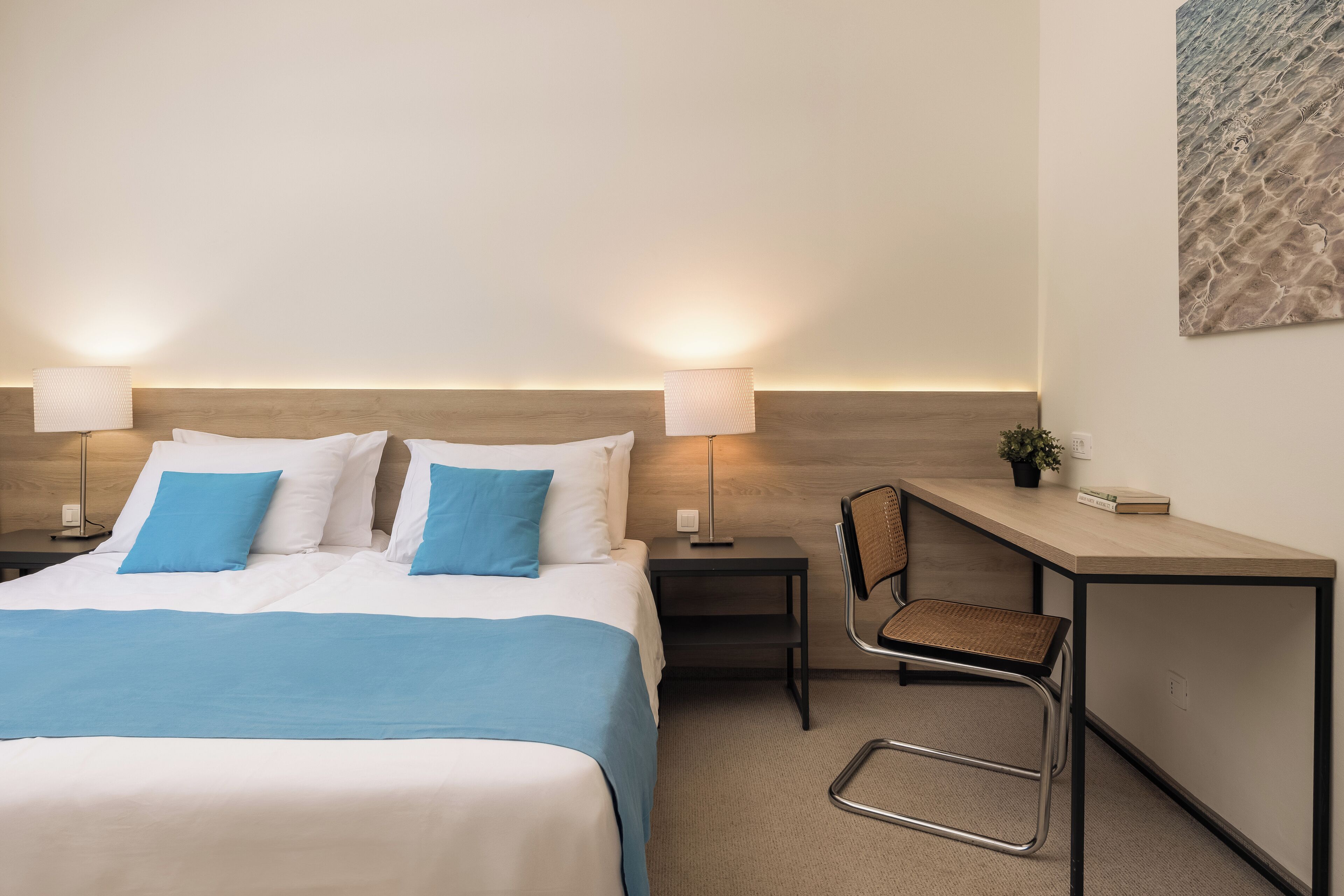 A hotel room with a bed, two bedside lamps, a desk with a chair, and a wall-mounted artwork.