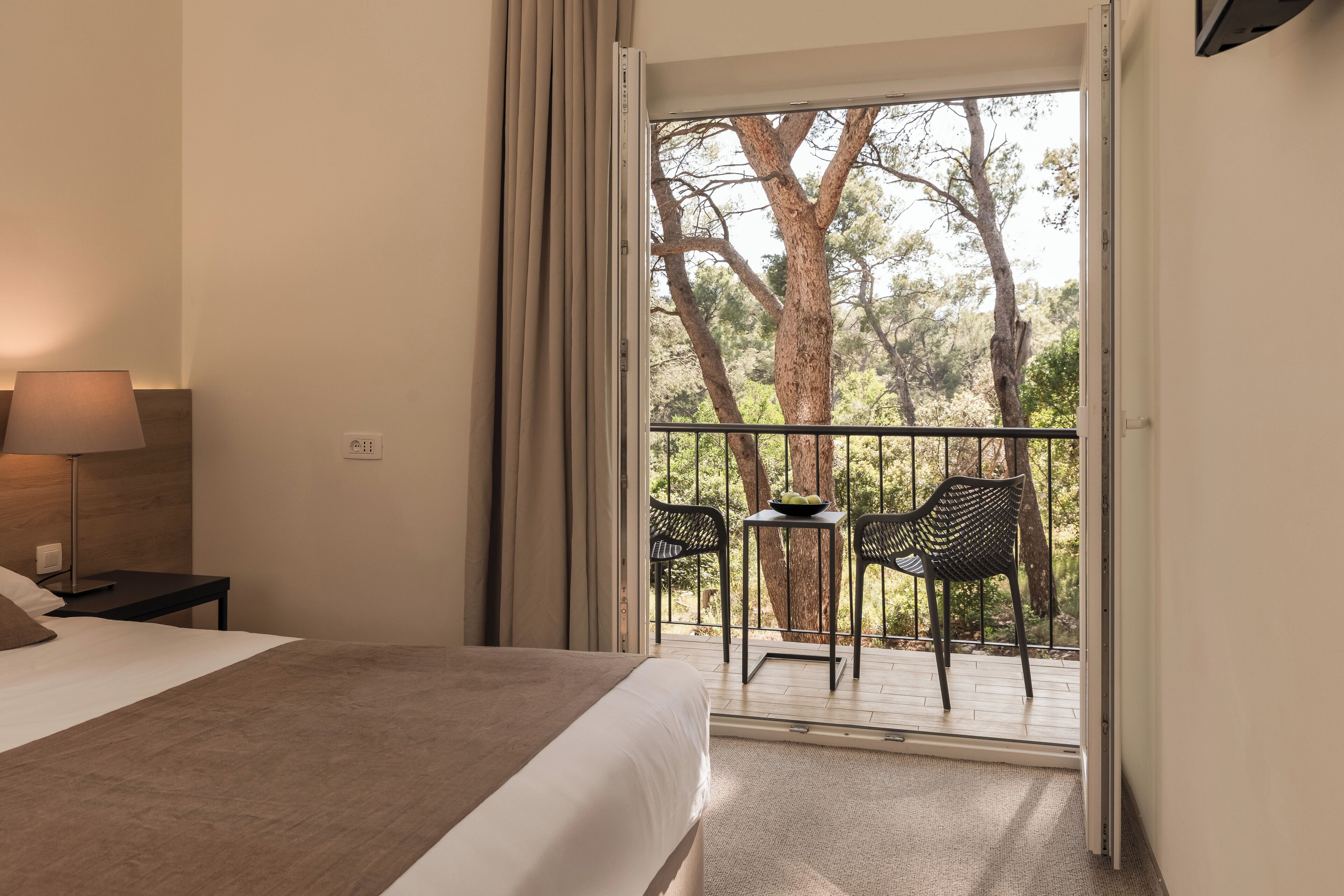 Un dormitorio con cama, mesita de noche, lámpara y vistas a un balcón exterior con sillas y una mesa.