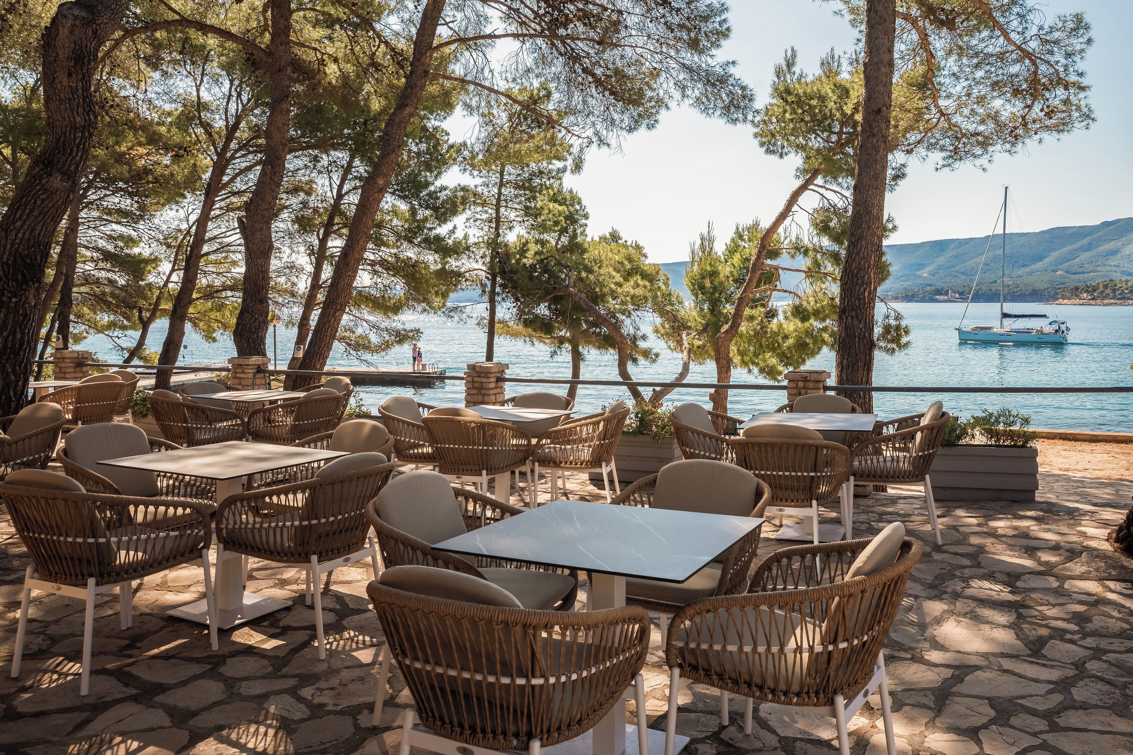 Een terras met tafels en rieten stoelen, uitkijkend op een water met een zeilboot.