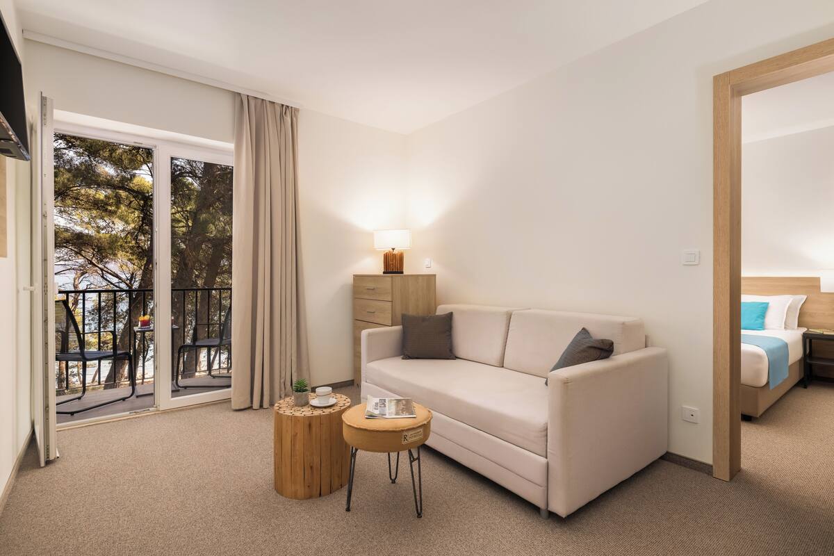 A modern hotel room with a sofa, a small round table, a lamp, and a view of the outdoors through a sliding door.