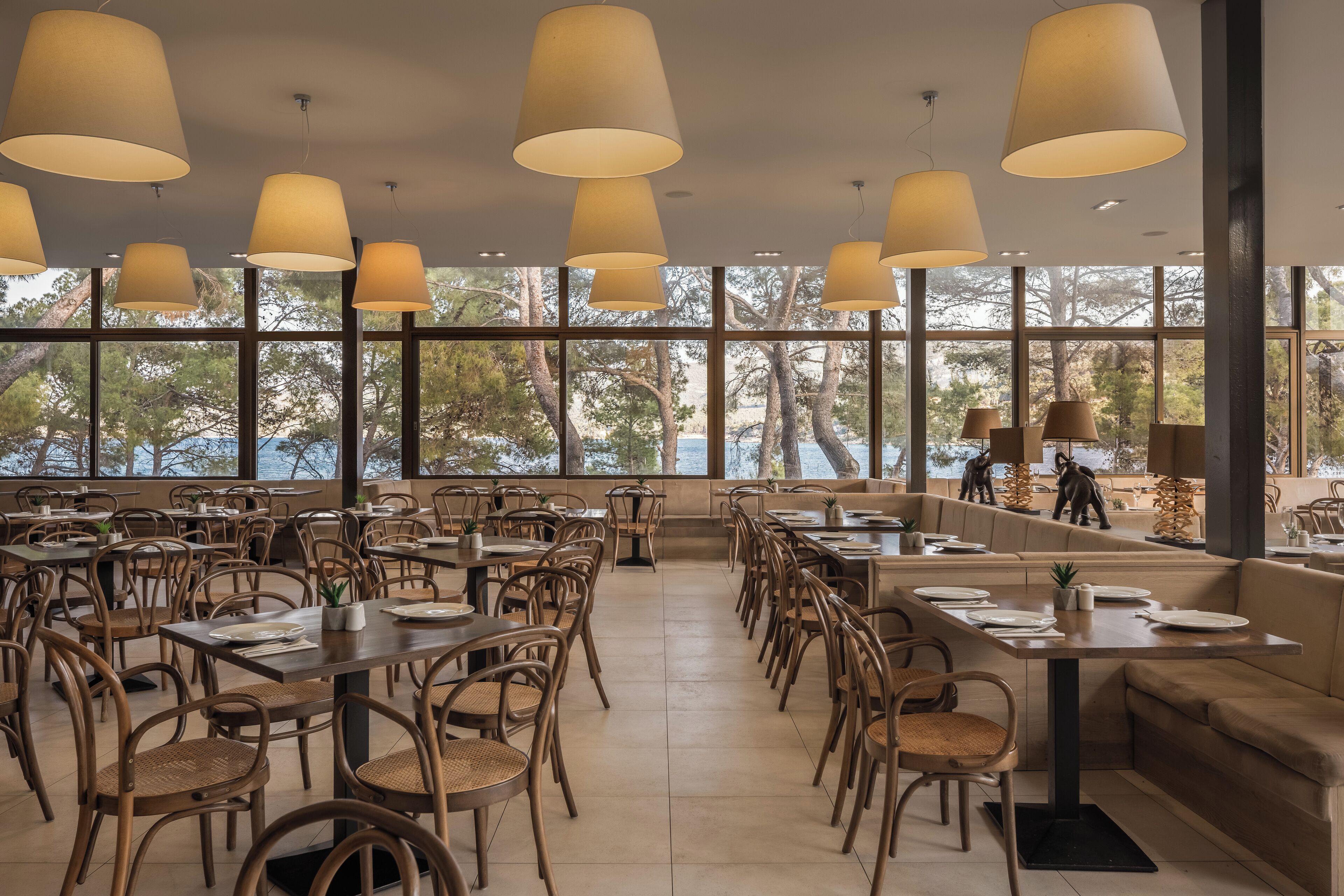 A spacious restaurant with high ceilings, large windows, and a view of trees and a body of water. Tables are set with white tablecloths and chairs.