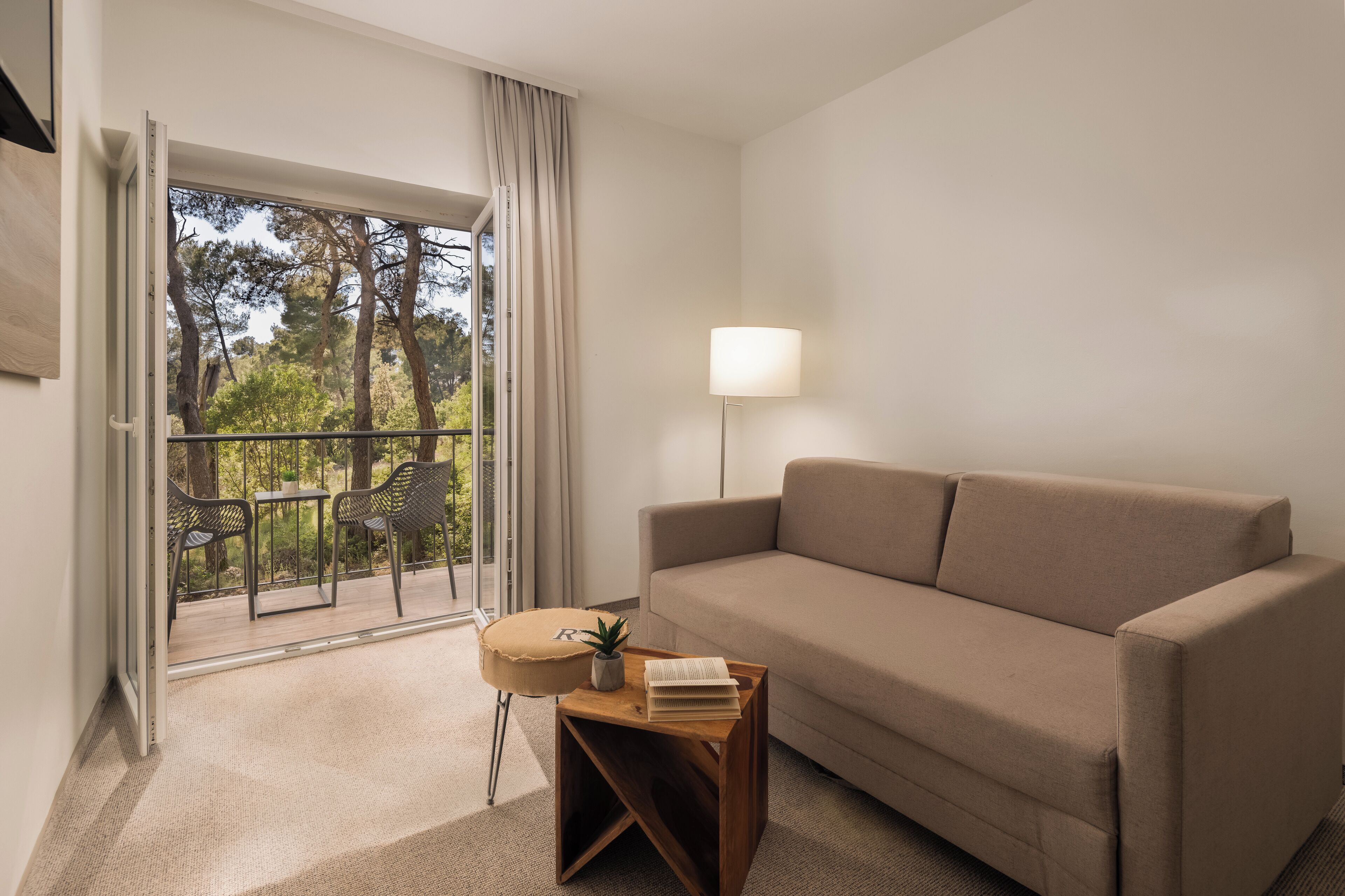 A modern living room with a beige sofa, a small wooden side table with a lamp, and a view of a balcony with outdoor furniture.