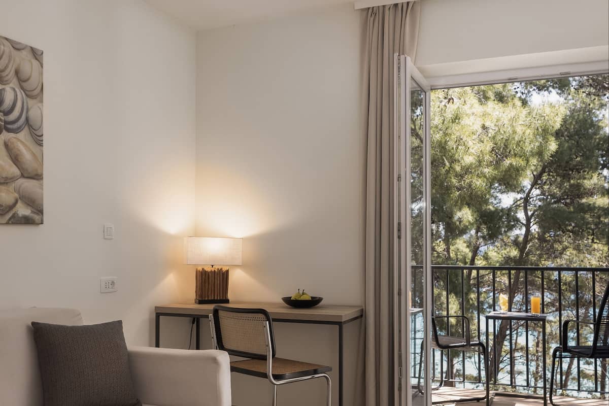 A hotel room with a dining area, a table with chairs, a lamp, and a view of trees through the balcony doors.