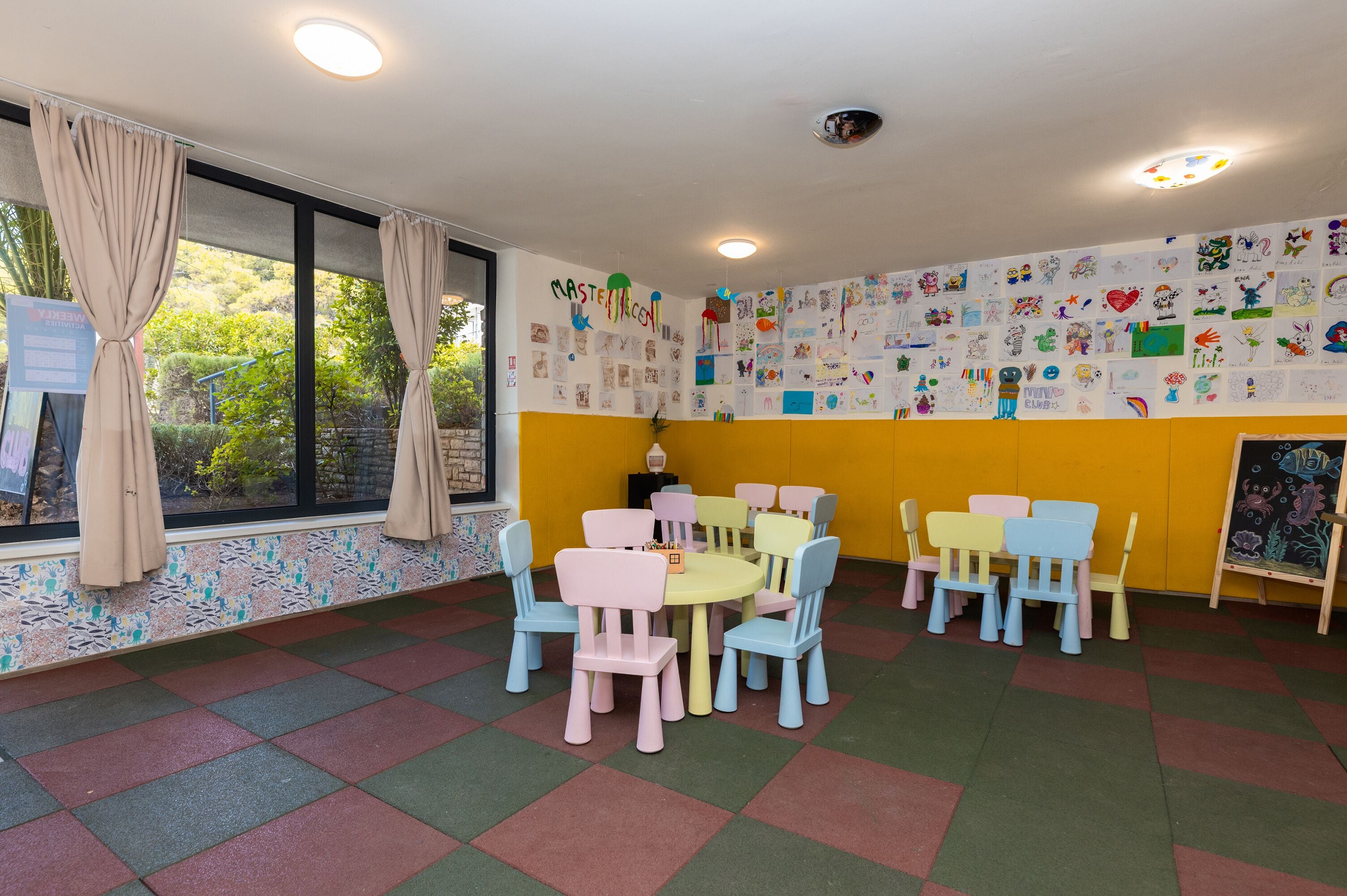 A children's playroom with colorful chairs and tables, decorated walls with children's drawings, and large windows with curtains.