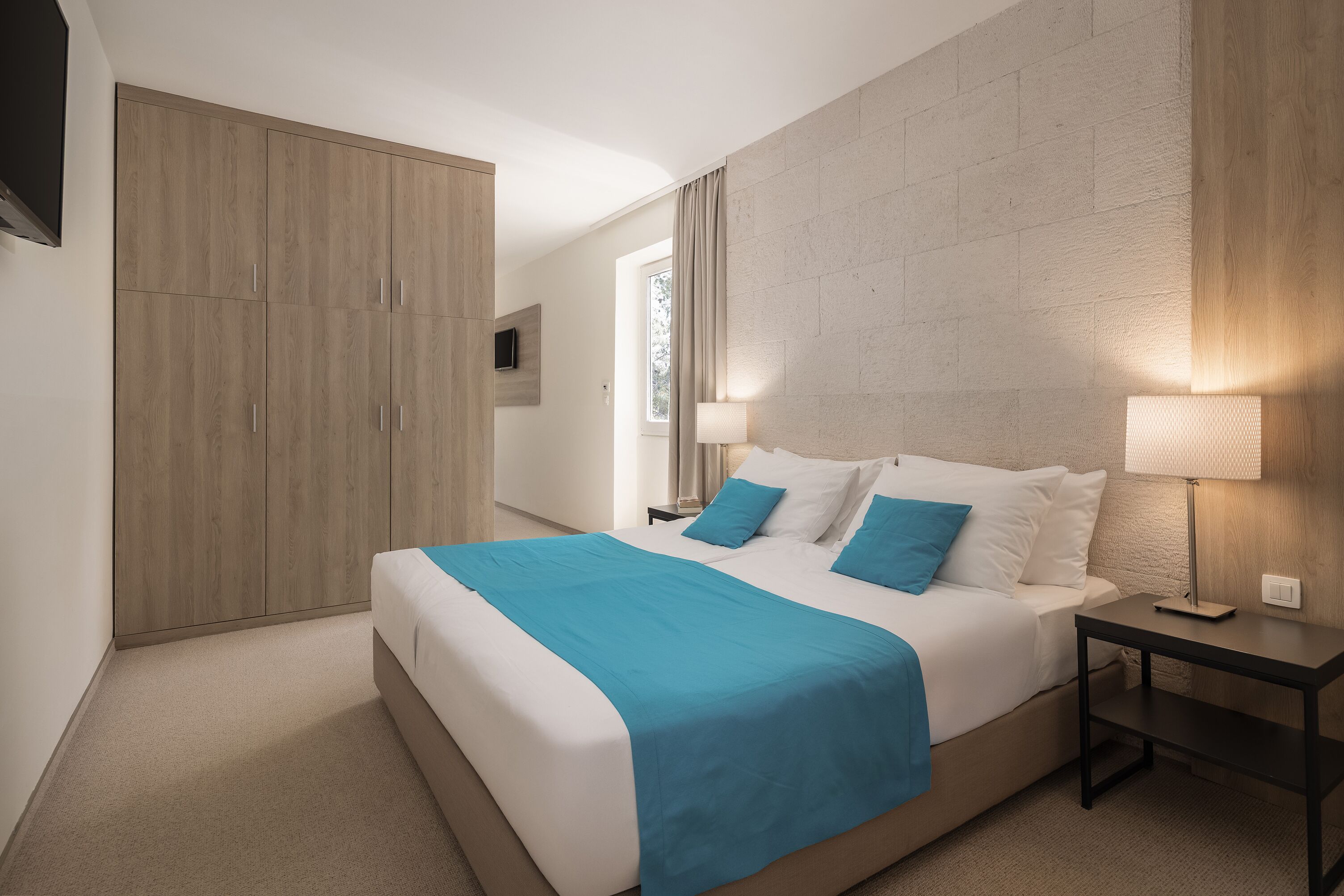 A modern hotel room with a large bed, a bedside table with a lamp, a wardrobe, and a television mounted on the wall.