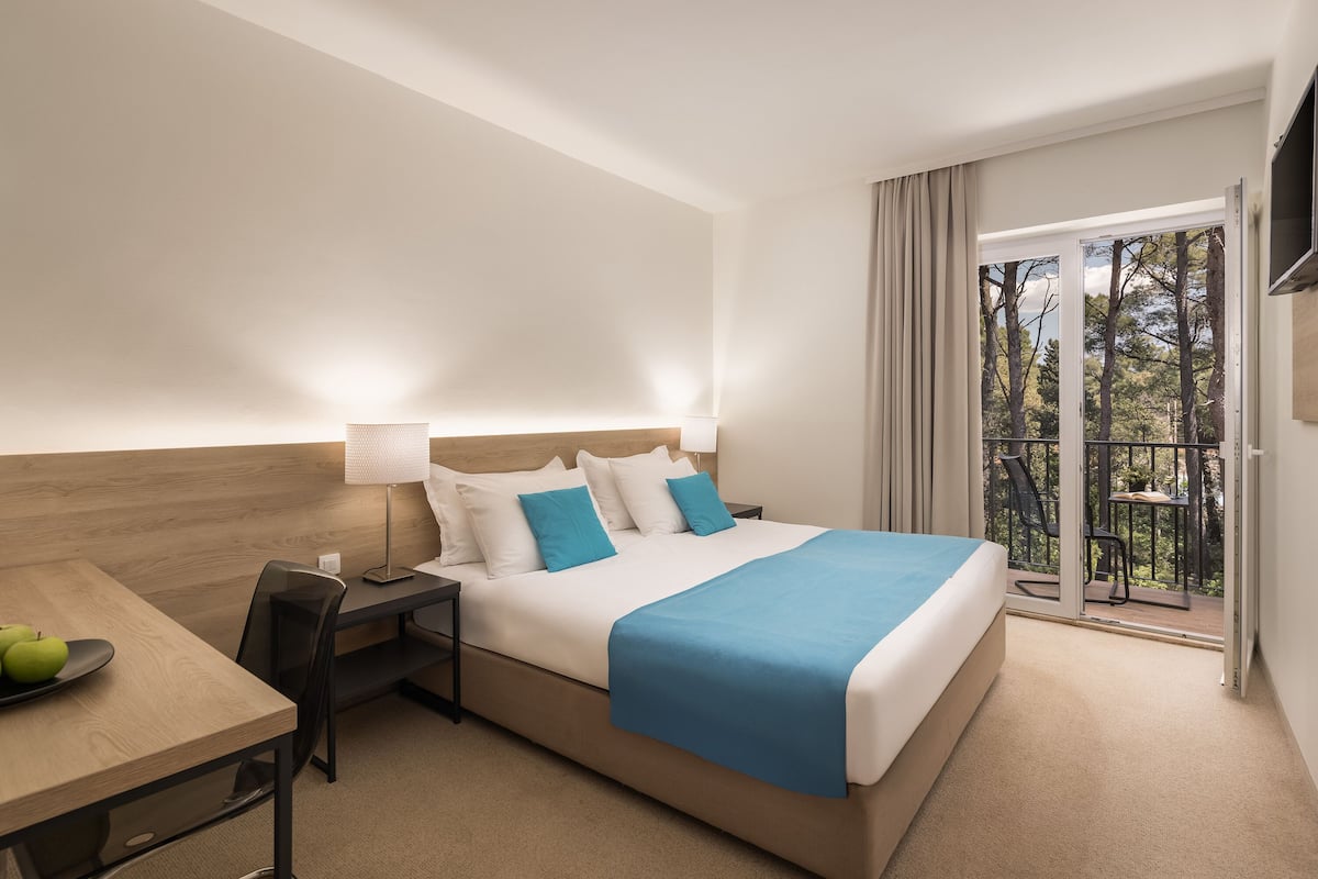 A hotel room with a bed, a desk with a chair, a laptop, and a view of the outdoors through a sliding door.
