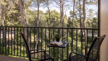 A balcony with two black metal chairs, a small table with an open book and a glass of water, and a view of a forested area.