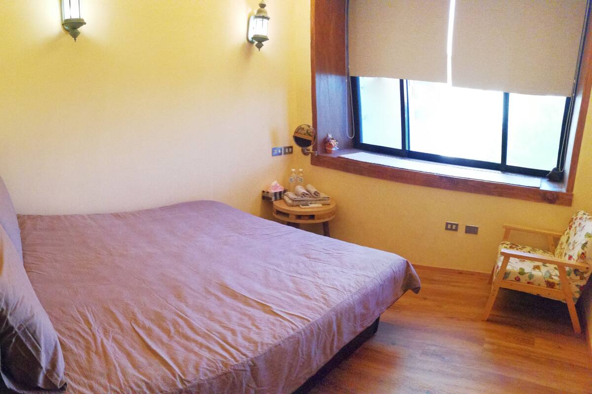 A bedroom with a bed, a chair, a small table, a window with blinds, and a wall-mounted light fixture.