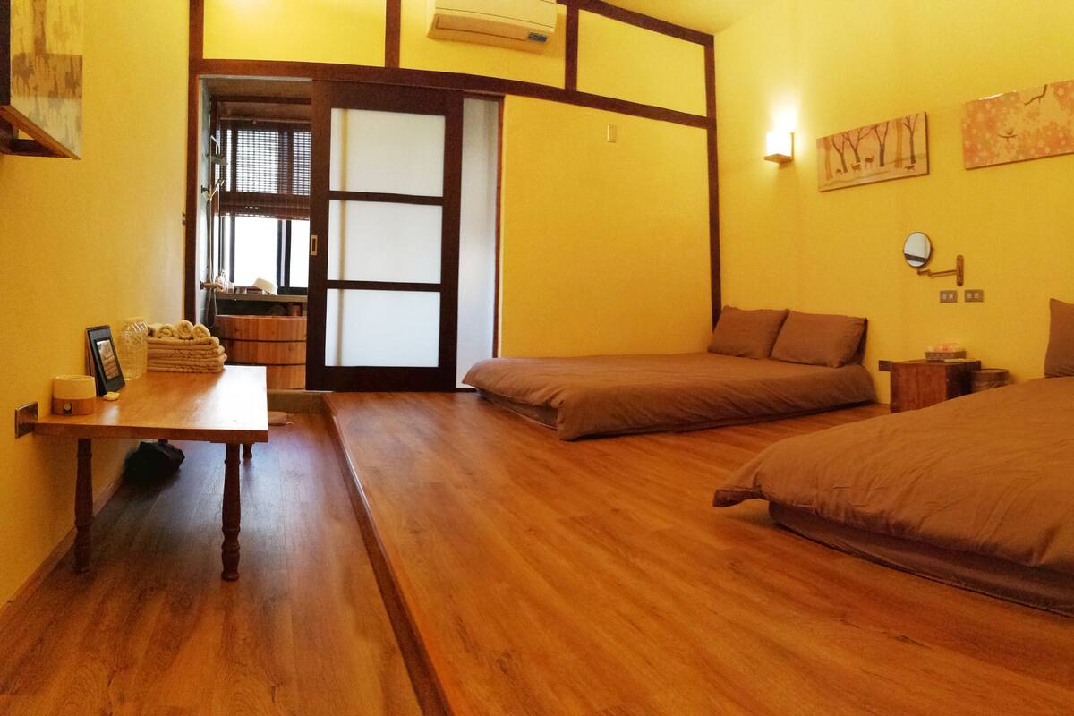 A room with a wooden floor, two single beds, a small table, and a sliding door leading to another room.