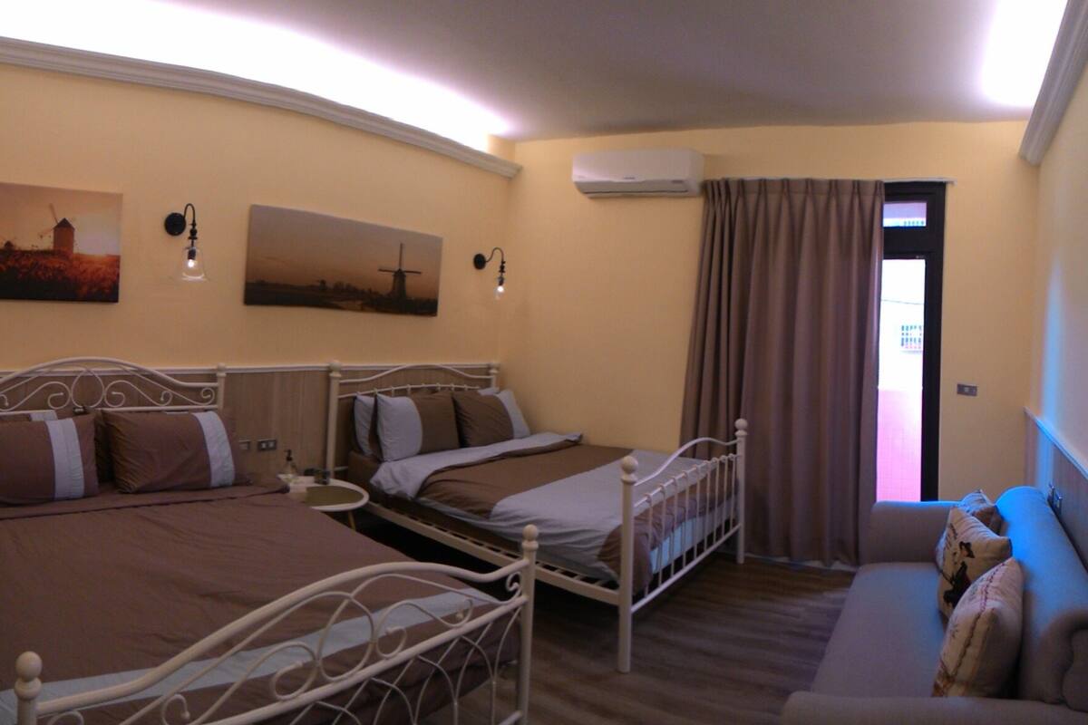 A hotel room with two beds, a sofa, a painting, and an air conditioning unit.