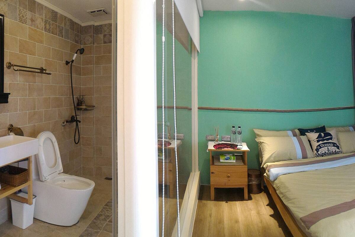 A hotel room with a shower, toilet, bedside table, and a bed with pillows and a blanket.