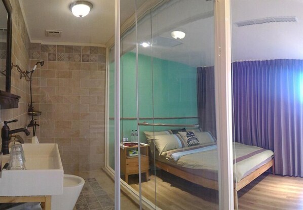 A hotel room with a large bed, a bedside table, a mirror, a sink, and a shower area.