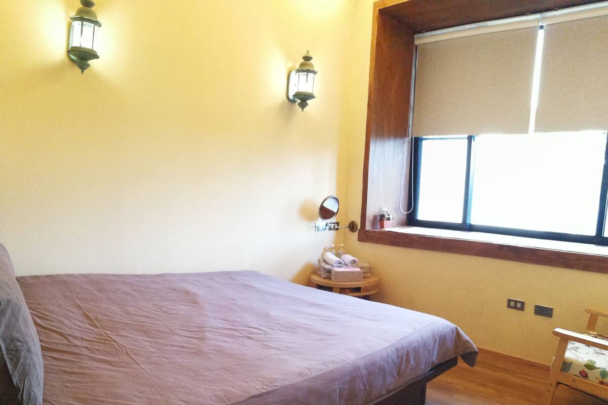 A bedroom with a bed, a window with blinds, a bedside table with a mirror, and a small wooden chair.