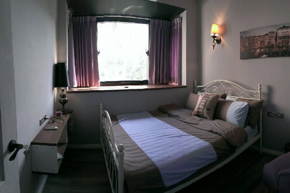 A bedroom with a single bed, a nightstand, a lamp, a window with curtains, a wall-mounted lamp, and a painting.
