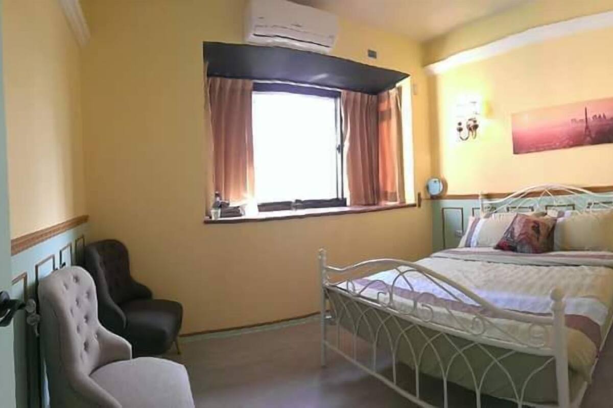 A bedroom with a single bed, a chair, a window with curtains, and a wall-mounted air conditioner.