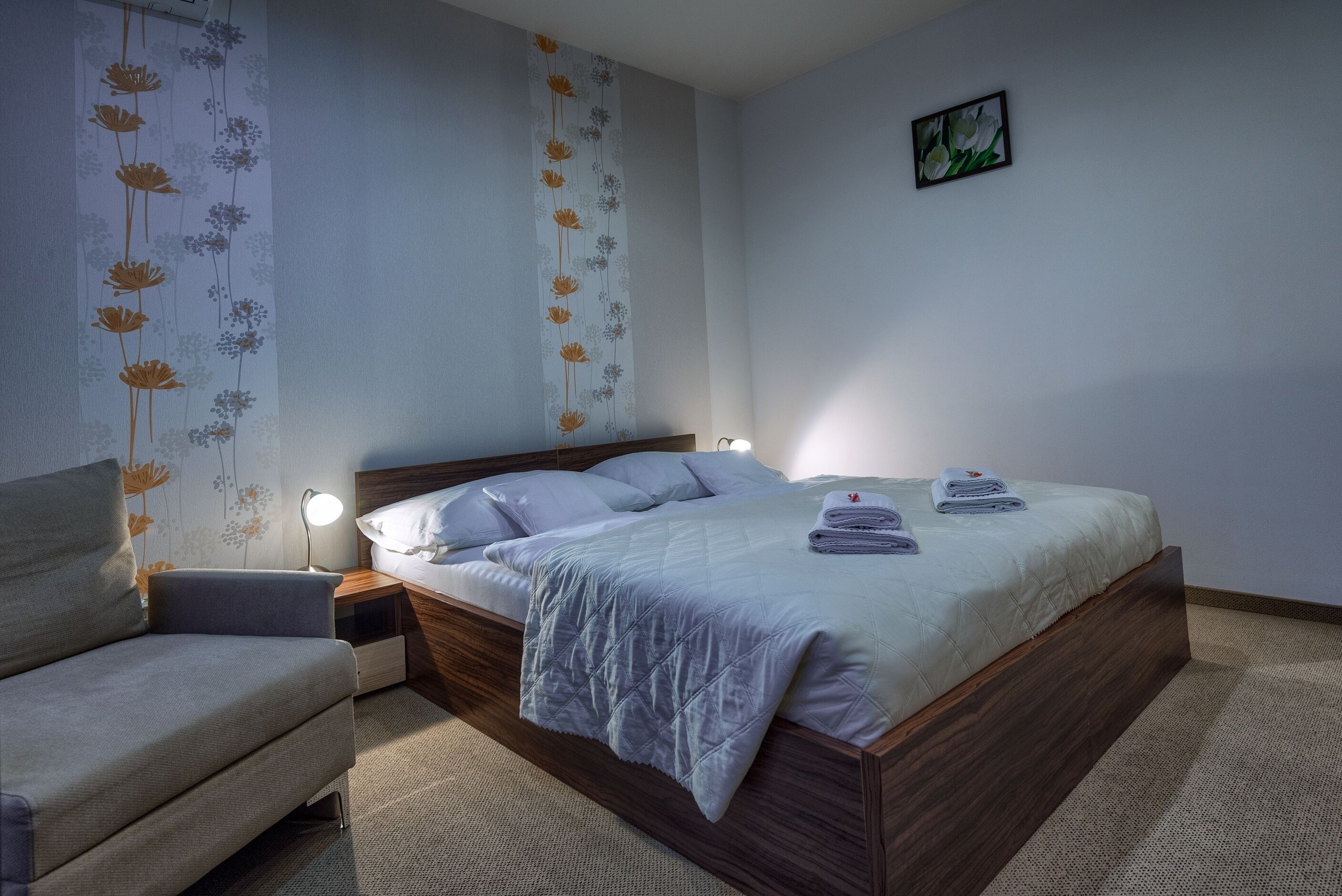Une chambre à coucher comprenant un lit, des tables de chevet, une chaise et un mur recouvert d’un papier peint à motifs floraux.