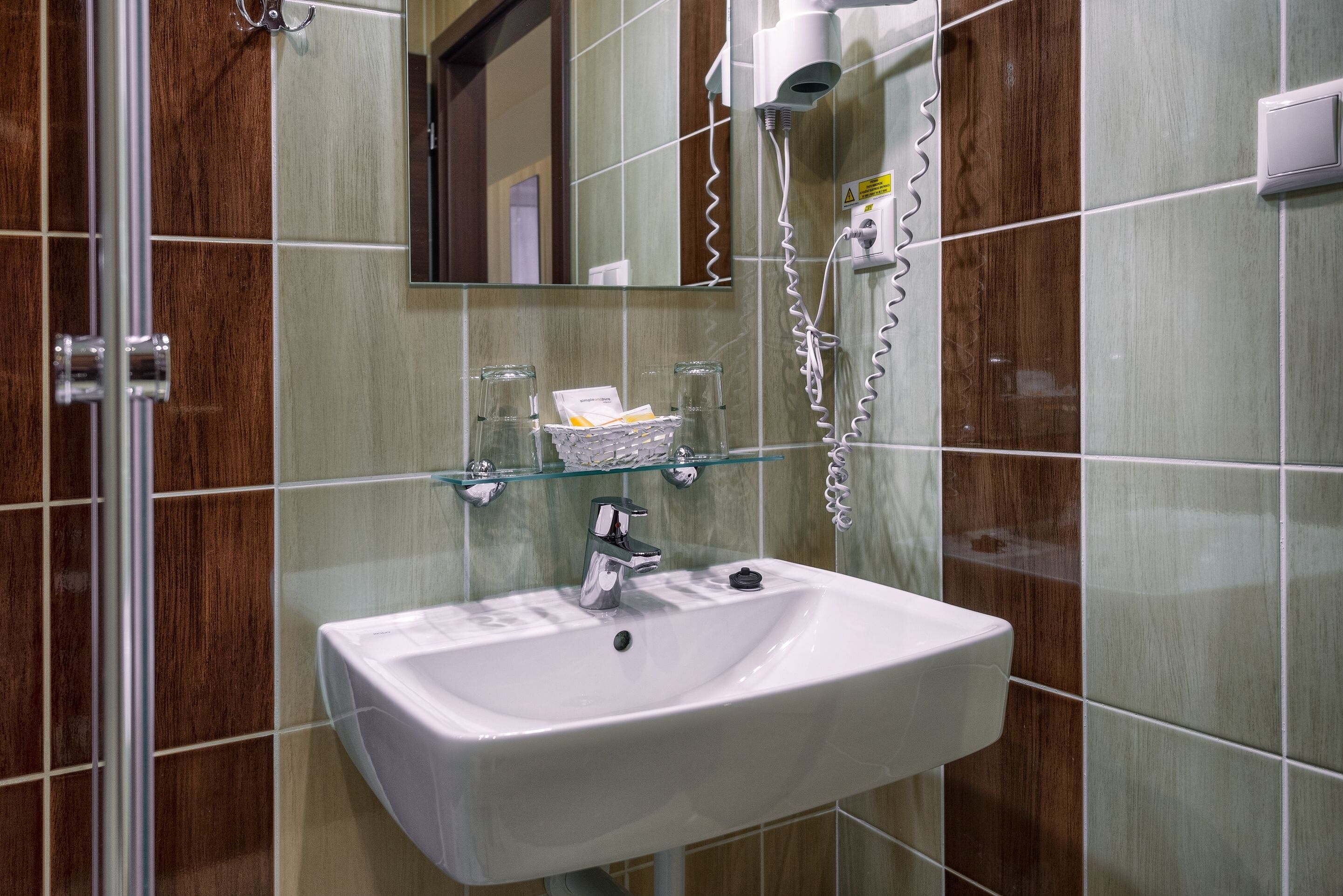 A bathroom with a white sink, a glass shelf above it, and a hairdryer on the wall.