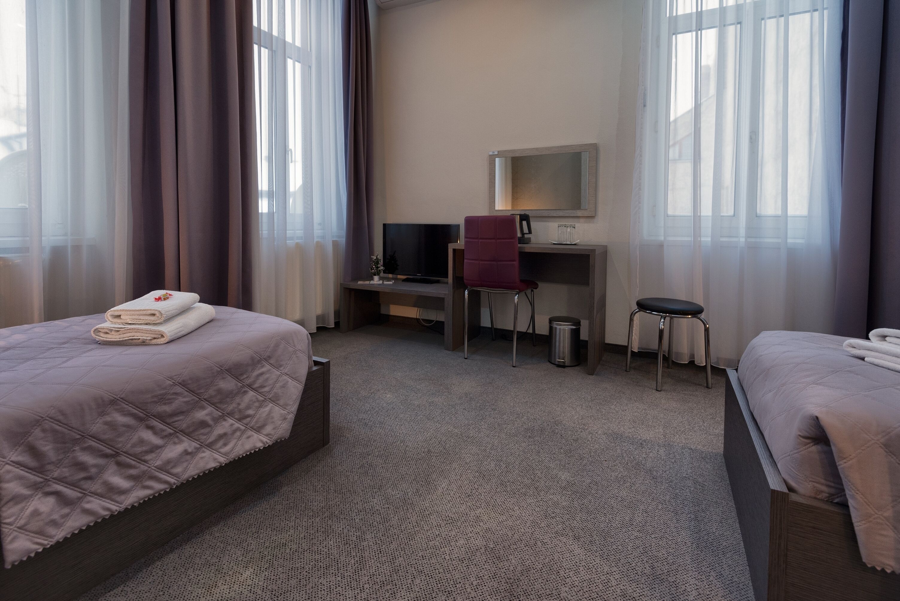 A hotel room with two beds, a desk, a chair, a TV, and a small stool.