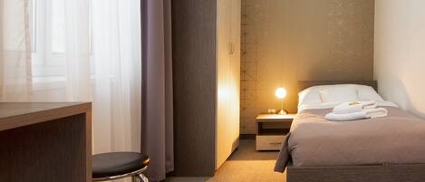 A hotel room with a bed, a desk, a chair, and a nightstand with a lamp.