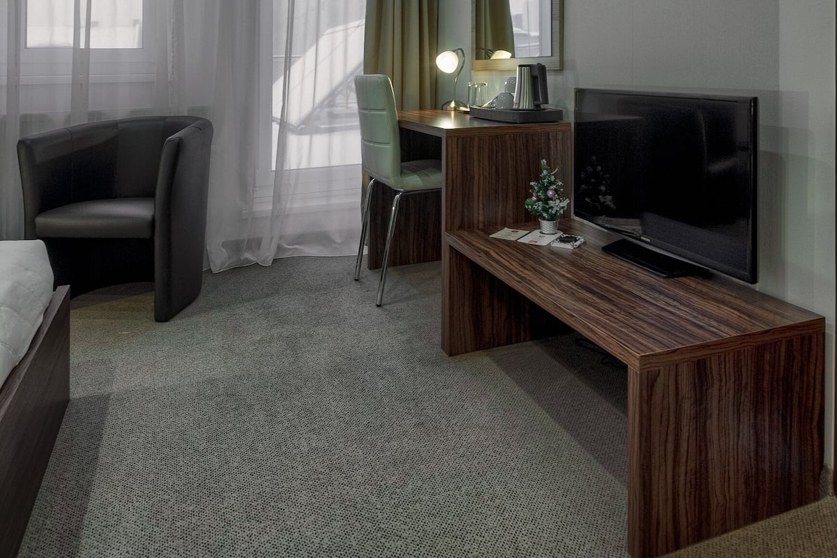 A hotel room with a bed, a desk with a computer, a chair, a TV, and a window with sheer curtains.