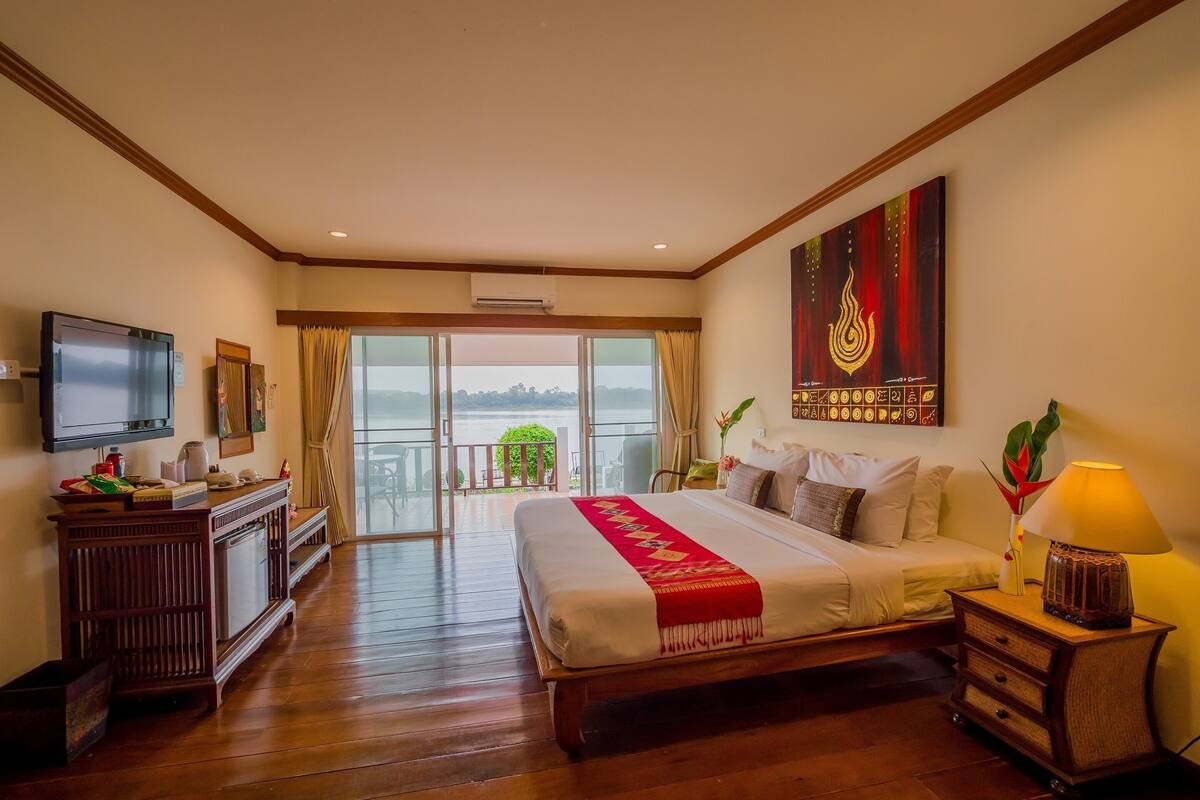A hotel room with a large bed, a TV, a desk, and a balcony with a view of the water.