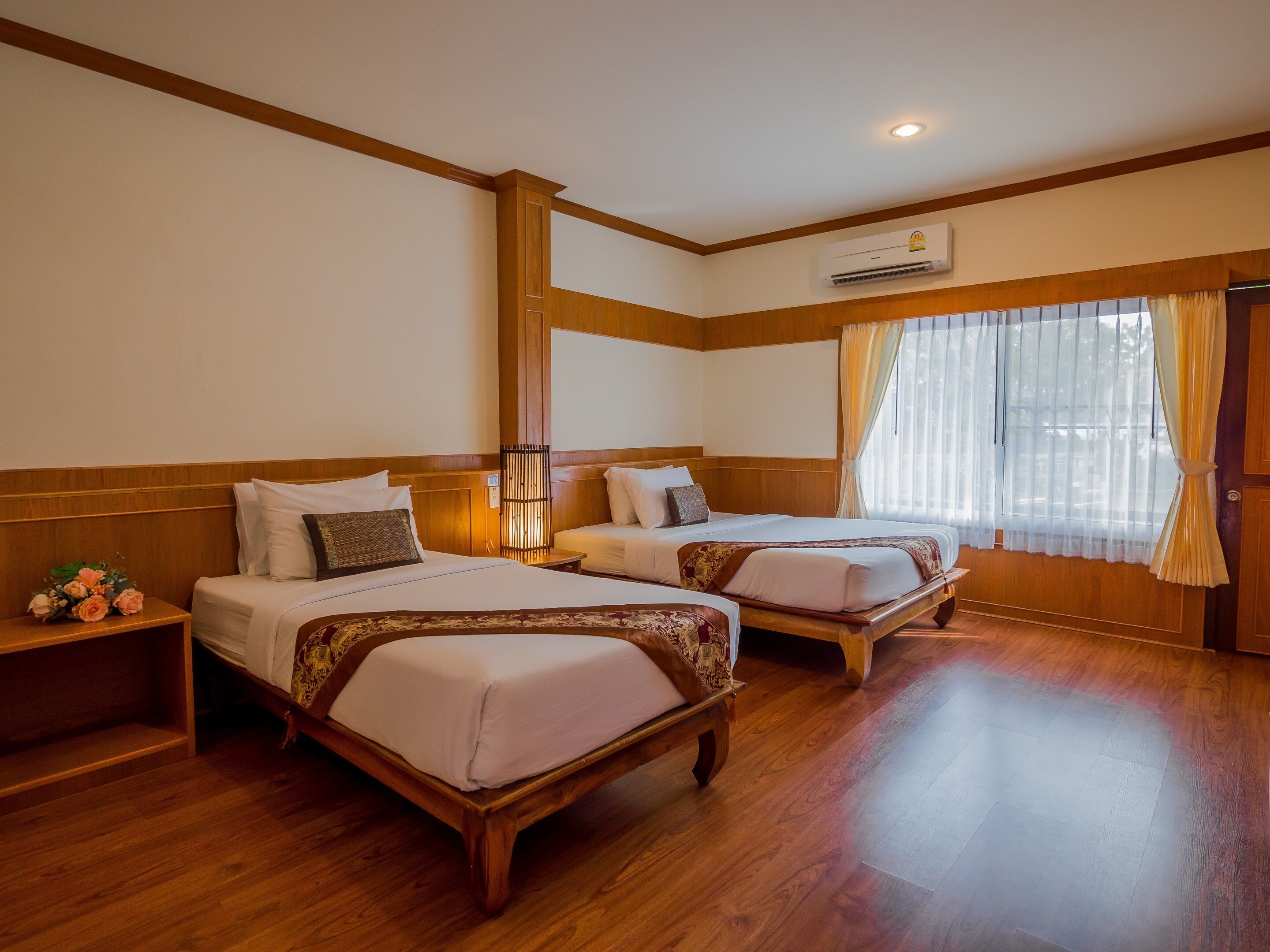 A hotel room with two wooden beds, a wooden floor, a window with curtains, and an air conditioning unit.