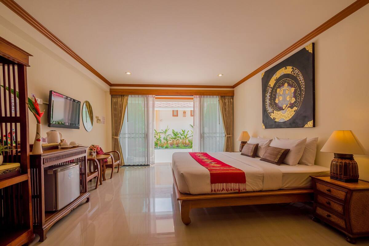 A hotel room with a bed, bedside table, lamp, TV, and a view of the outside.