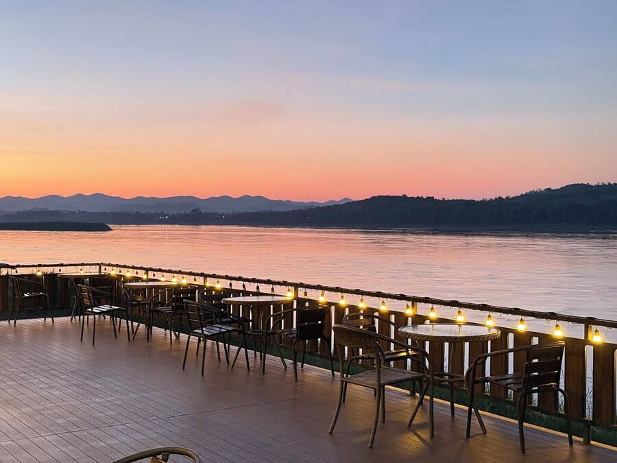 Terraza a la orilla del río con mesas y sillas, luces colgantes y vistas al río y colinas al lejano.