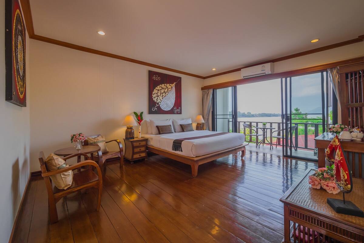 A spacious bedroom with a large bed, a wooden floor, a balcony with a view, and a dining area with chairs and a table.