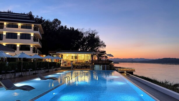 Una piscina con tumbonas y sombrillas, un restaurante con terraza y vistas a una masa de agua al atardecer.