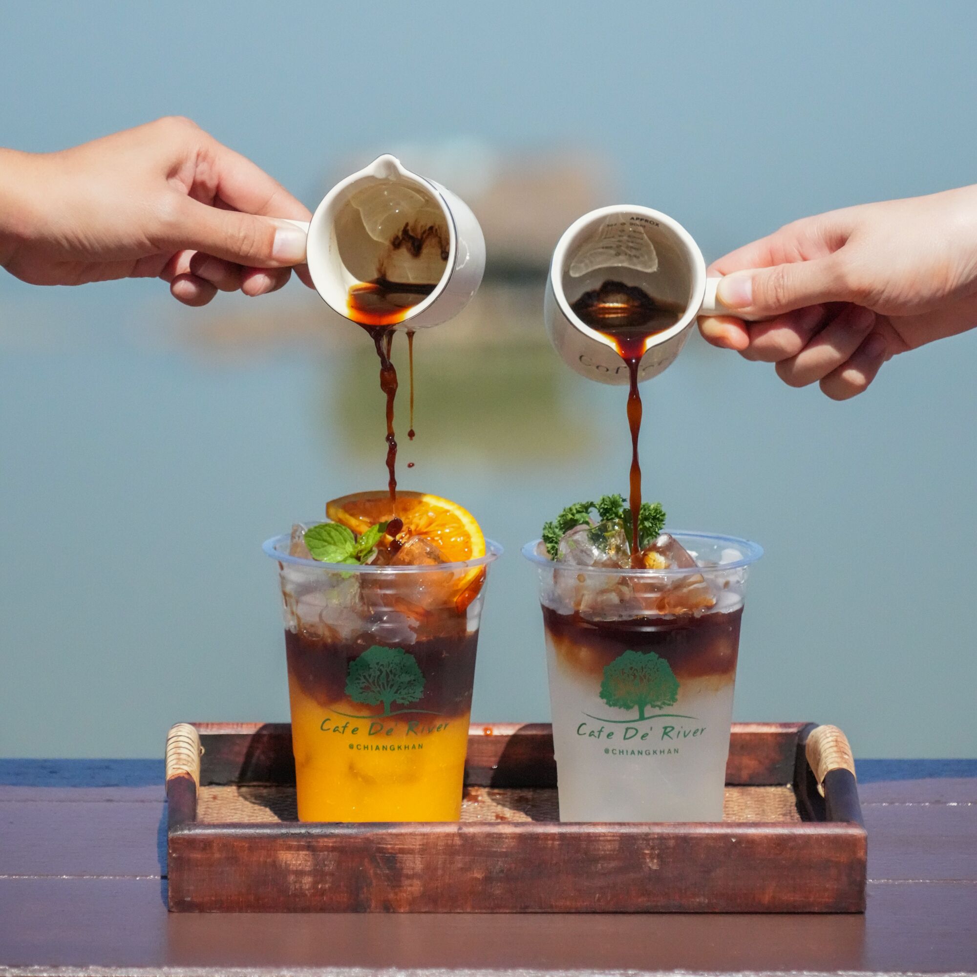 Twee ijskoude drankjes met een boomlogo en de tekst 'Cafe De River' op een dienblad.