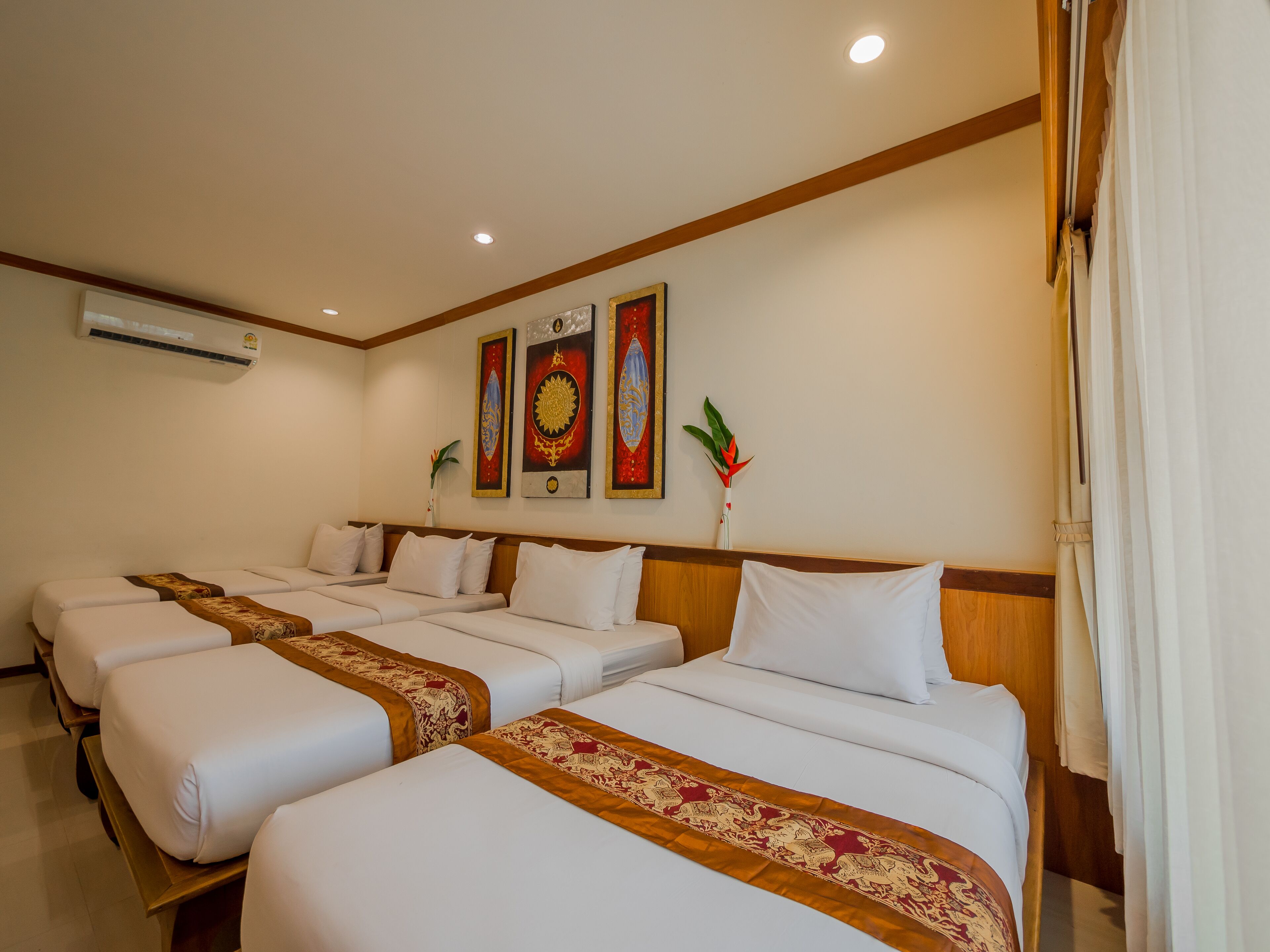 Habitación de hotel con tres camas individuales, cada una con colchón blanco y corredor de cama decorativo rojo y dorado. Se observan tres unidades de aire acondicionado instaladas en la pared, y un cuadro decorativo grande sobre las camas.