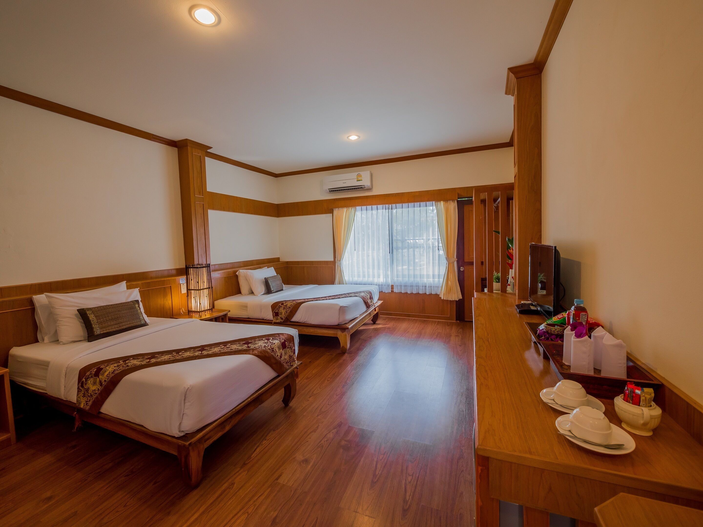 A hotel room with two beds, a wooden floor, a TV, and a small table with cups and a bowl.